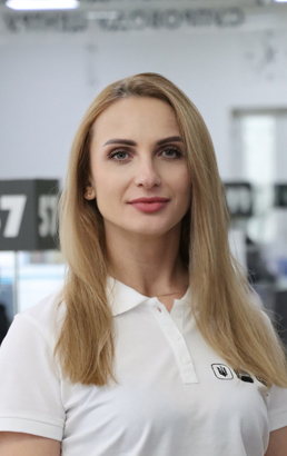 Молода жінка зі світло-русявим волоссям у білій футболці стоїть в офісі.